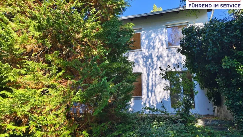 Das Haus ist von üppigem Grün umgeben und zeigt eine schlichte Fassade mit mehreren Fenstern.