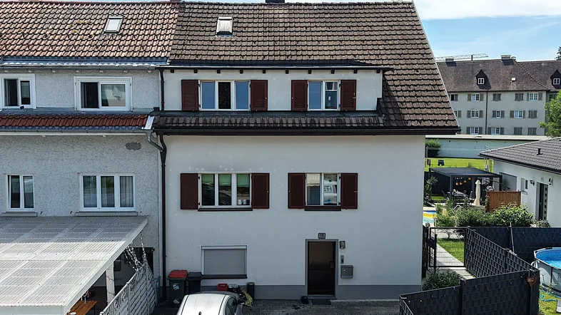 Außenansicht eines gepflegten Hauses mit Satteldach, braunen Fensterläden, Einfahrt und einem Gartenbereich mit Pool.