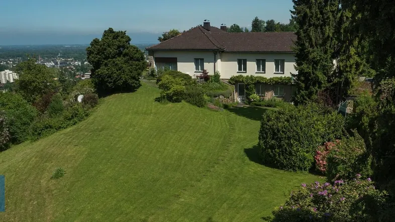 Luftaufnahme eines Hauses auf einem Hügel mit weitläufigem Rasen und Panoramablick auf die Stadt.