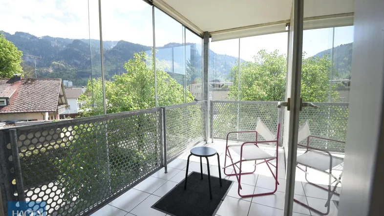 Verglaster Balkon mit Sitzgelegenheiten und Panoramablick auf die umliegenden Berge und grüne Bäume.