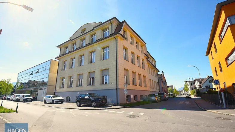 Großes Eckgebäude mit klassischer Architektur und modernen Anbauten an einer sonnigen Straße.