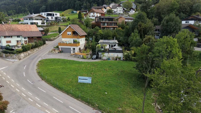 Luftaufnahme eines Baugrundstücks in bergiger Umgebung mit umliegenden Häusern und Straßen.