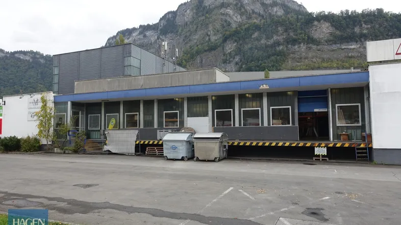 Gebäudeansicht eines Gewerbeobjekts mit Laderampe und Müllcontainern vor einer Bergkulisse.