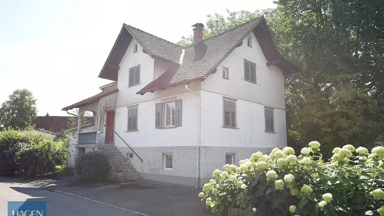 Weißes Einfamilienhaus mit Satteldach, grünen Fensterläden und gepflegter Vorgartenbepflanzung unter strahlendem Himmel.