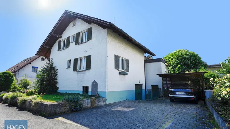 Weißes Einfamilienhaus mit grünen Fensterläden, gepflasterter Auffahrt und Carport.