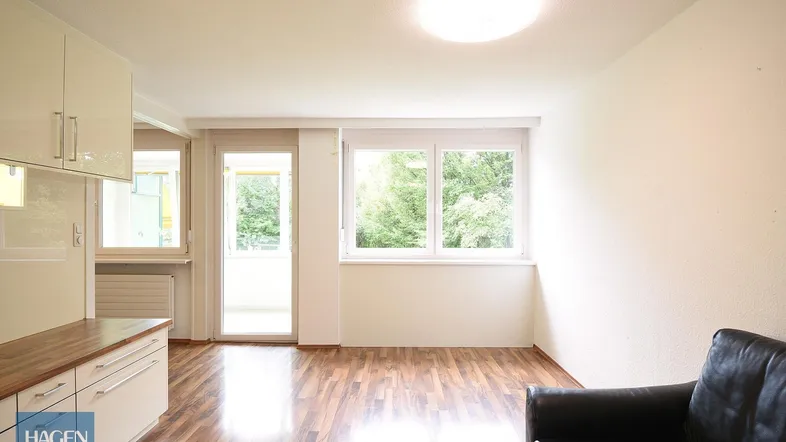 Heller Wohnbereich mit Holzboden, großen Fenstern und Zugang zu einem Balkon mit Grünblick.