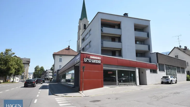 Außenansicht eines modernen Gebäudes mit Fahrradgeschäft im Erdgeschoss und Wohnungen darüber, im Hintergrund eine Kirche.