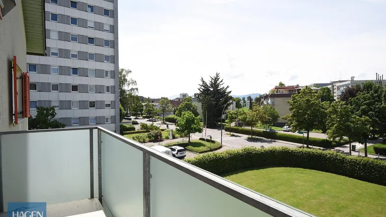 Blick vom Balkon auf die grüne Umgebung und umliegende Gebäude mit einem Hochhaus im Hintergrund.