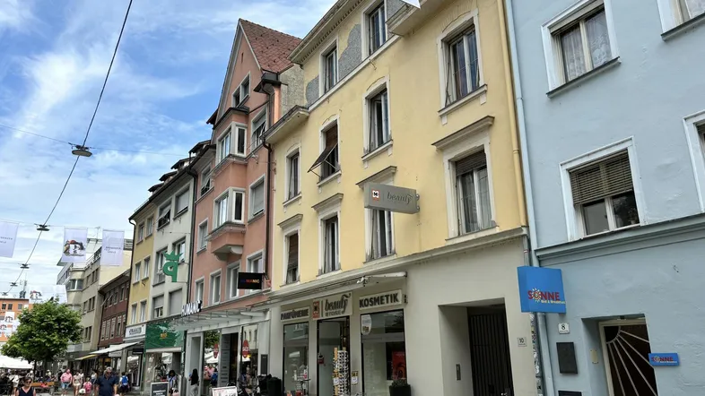 Seitenansicht eines gelben Mehrfamilienhauses mit Geschäften und belebter Fußgängerzone.