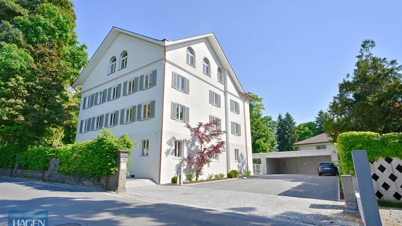 Weißes Mehrfamilienhaus mit grünen Fensterläden, umgeben von Bäumen und einem gepflegten Vorgarten mit Zufahrt.