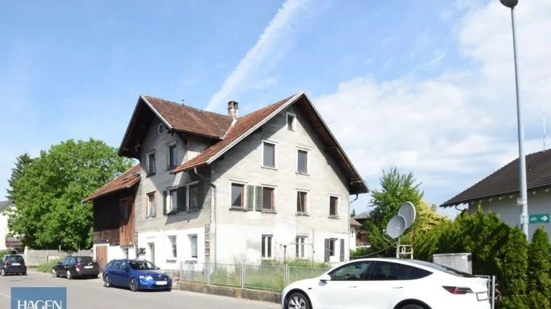 Altes Wohnhaus mit Satteldach an einer Straße, umgeben von Bäumen und parkenden Autos.