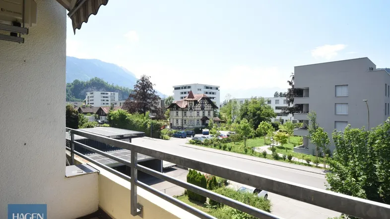 Balkon mit Blick auf die Straße, umliegende Gebäude und ferne Berge unter blauem Himmel.