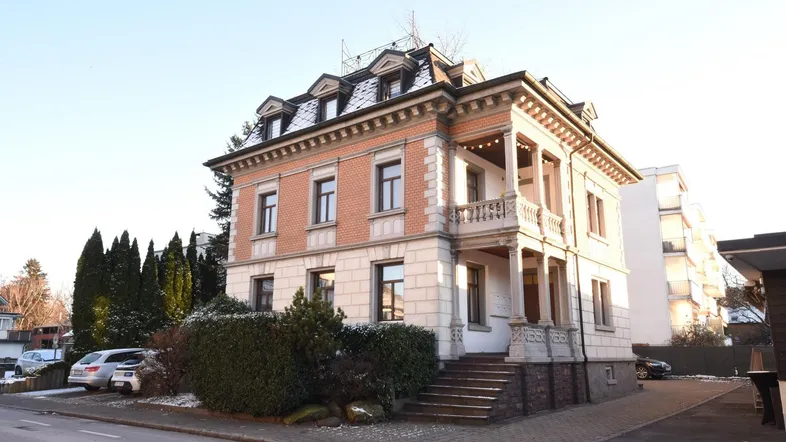 Historisches Gebäude mit klassischer Architektur, Mansarddach und Balkonen, umgeben von winterlicher Vegetation.