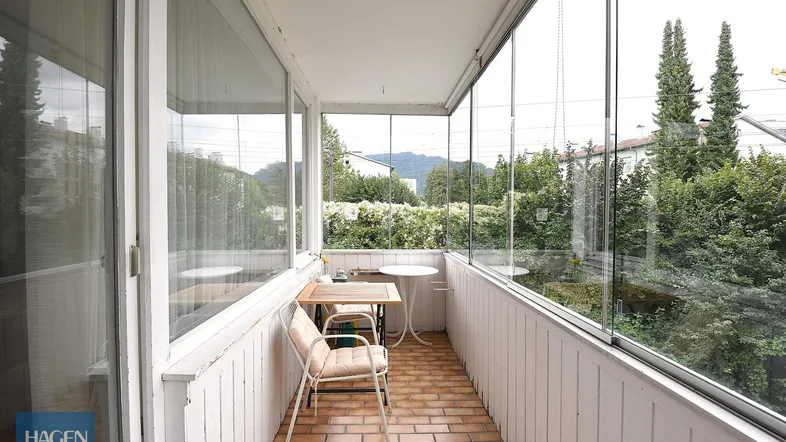 Verglaster Balkon mit Fliesenboden, Sitzgelegenheiten und Blick auf Bäume und Berge.