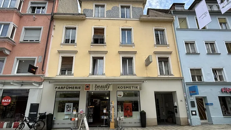 Gelbe Fassade eines Mehrfamilienhauses mit Geschäften im Erdgeschoss an einer belebten Straße.
