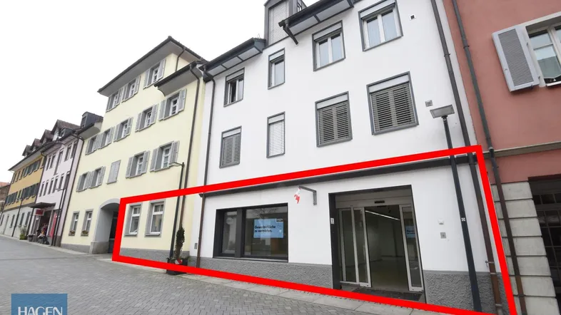Außenansicht eines Geschäftsgebäudes mit Schaufenstern und Eingang an einer gepflasterten Straße.