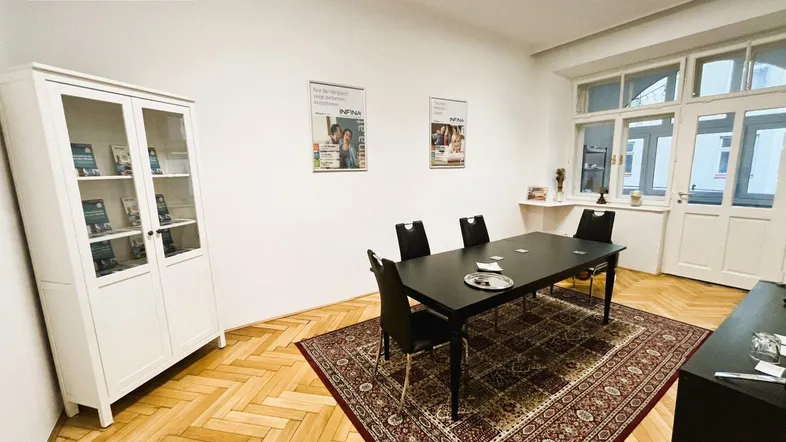 Heller Büroraum mit Fischgrätparkett, einem großen Tisch und einem Vitrinenschrank am Fenster.