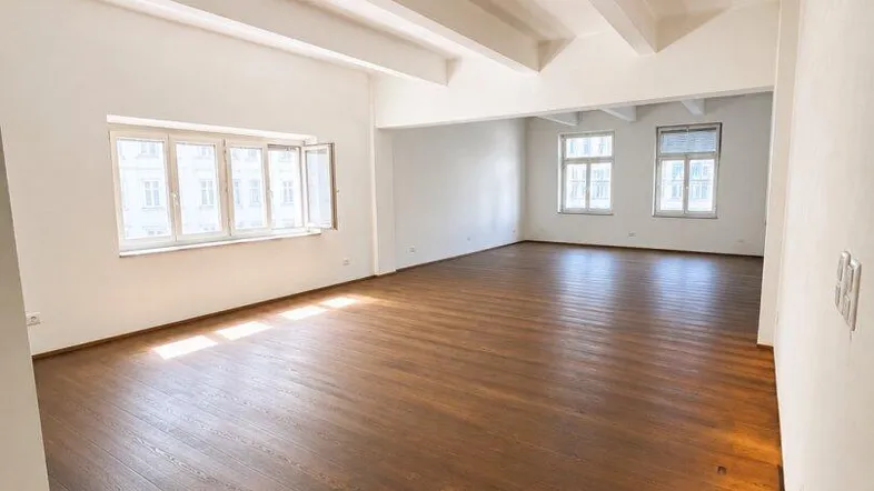 Geräumiger, leerer Raum mit dunklem Holzboden, weißen Wänden und sichtbaren Deckenbalken, ideal für flexible Nutzung.