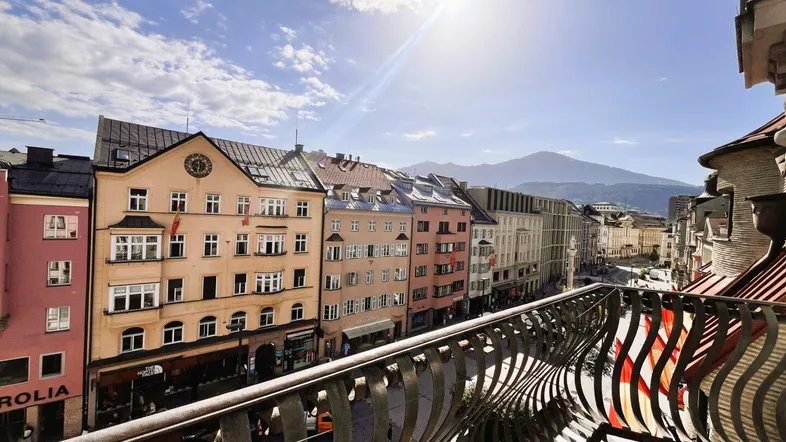 Sonniger Balkon mit schmiedeeisernem Geländer und Blick auf die historische Stadt und die Berge.
