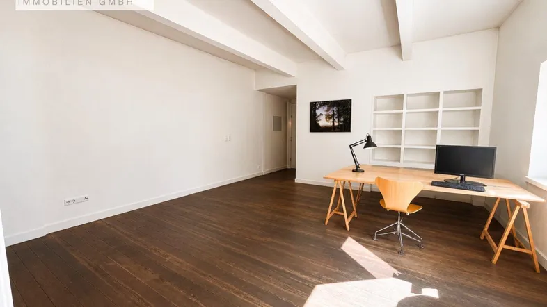 Geräumiger Raum mit dunklem Holzboden, weißen Deckenbalken und einem Arbeitsbereich mit eingebautem Regal.