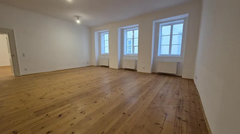Helles und geräumiges Zimmer mit Holzdielenboden und mehreren Fenstern, ideal als Wohnbereich.