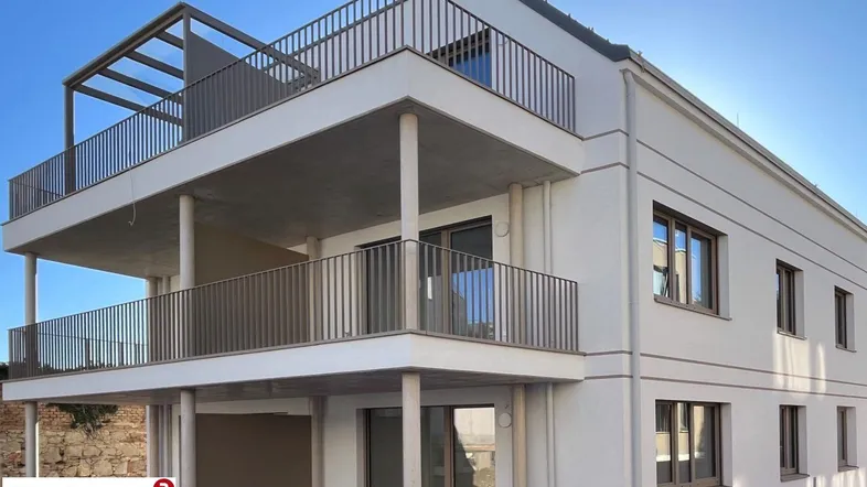 Modernes Mehrfamilienhaus mit mehreren Balkonen und einer Dachterrasse unter blauem Himmel.