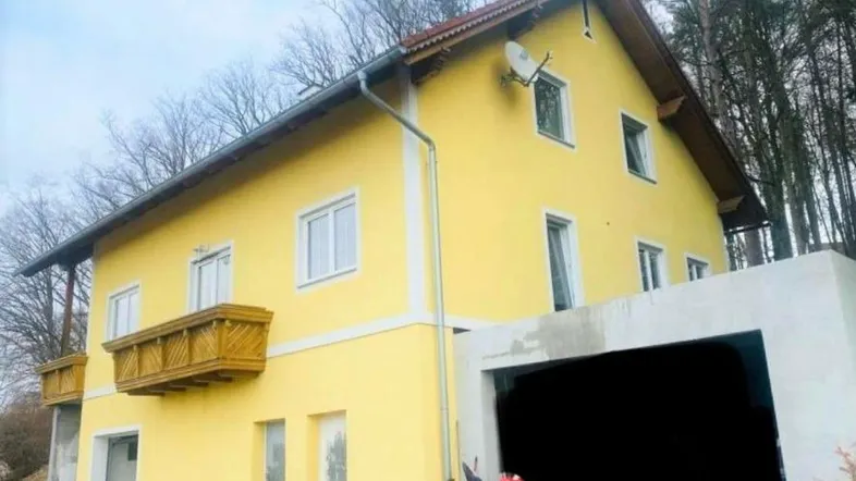 Gelbes Einfamilienhaus mit Holzbalkon und angrenzender Garage, umgeben von Bäumen.