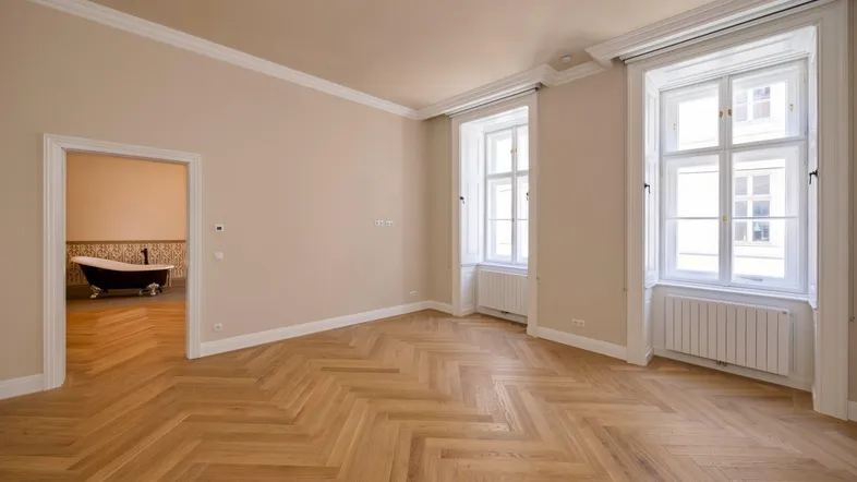 Geräumiges Zimmer mit Fischgrätparkett, hohen Decken und Stuckverzierung, mit Blick in ein angrenzendes Badezimmer.