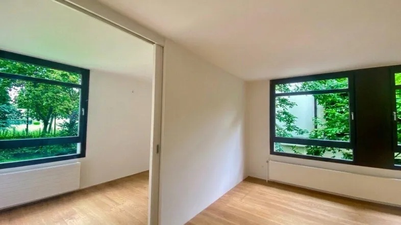 Helles Zimmer mit Holzboden und großen Fenstern, die einen Blick ins Grüne bieten.