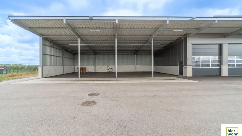 Große offene Lagerhalle mit Metallkonstruktion und asphaltiertem Vorplatz unter bewölktem Himmel.