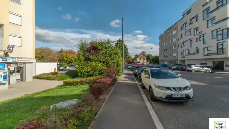 Blick auf die Straße und umliegende Gebäude mit parkenden Autos und Grünflächen unter blauem Himmel.
