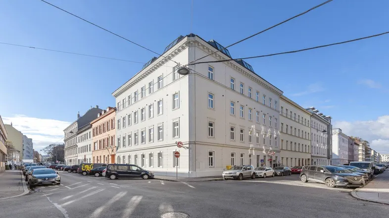 Eckgebäude in urbaner Umgebung mit klassischer Architektur und vielen Fenstern an einem sonnigen Tag.