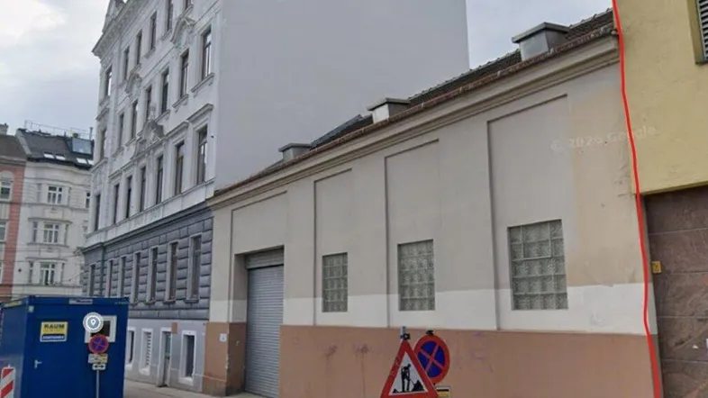 Straßenansicht eines Gebäudes mit schlichter Fassade, angrenzendem Baucontainer und Verkehrsschildern in urbaner Umgebung.