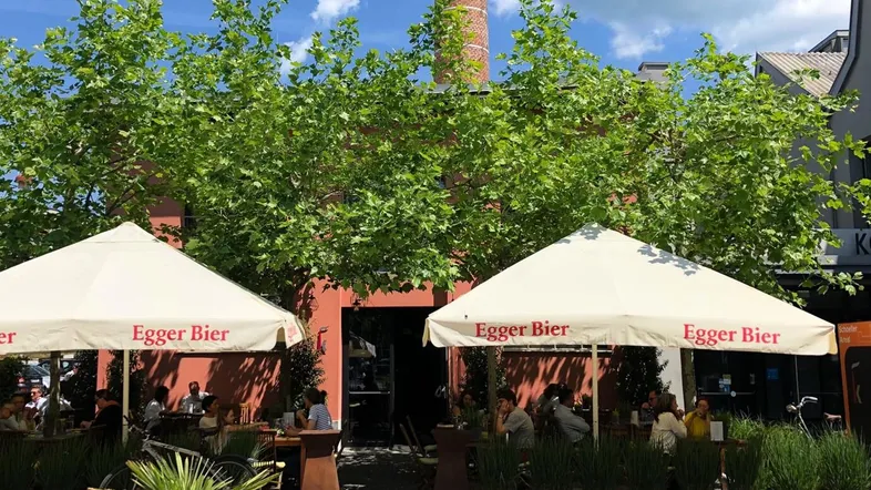 Gemütlicher Außenbereich mit Sonnenschirmen und Bäumen vor einem historischen Gebäude mit hohem Schornstein.