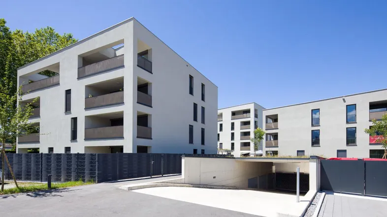 Moderne Wohngebäude mit weißer Fassade, Balkonen und Zufahrt zu einer Tiefgarage unter blauem Himmel.