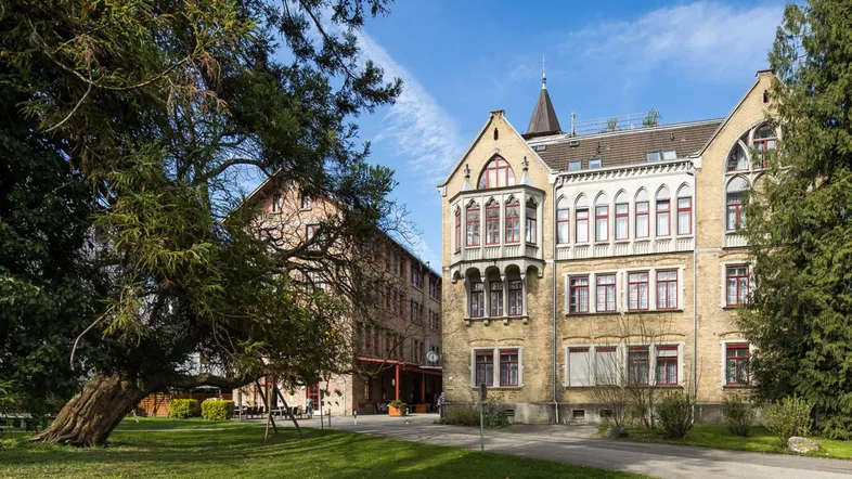 Zwei historische Gebäude am Benger Areal mit einem alten Baum im Vordergrund und blauem Himmel