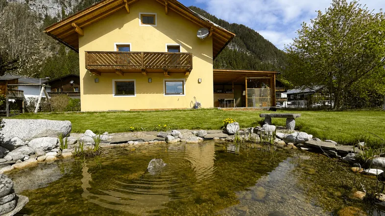 Gelbes Haus mit Holzbalkon, großem Garten, Teich und überdachtem Sitzbereich vor alpiner Bergkulisse.