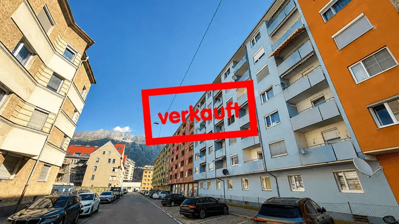 Mehrere Mehrfamilienhäuser entlang einer Straße mit geparkten Autos und Bergen im Hintergrund unter blauem Himmel.