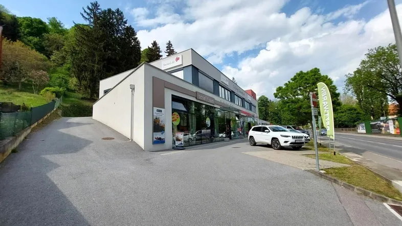 Modernes Geschäftsgebäude mit großen Schaufenstern und Parkplätzen an einer Straße, umgeben von grünen Hügeln.