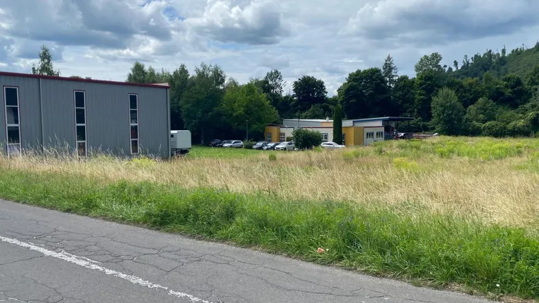 Großes, unbebautes Grundstück mit angrenzendem Gewerbegebäude und bewaldetem Hügel im Hintergrund.