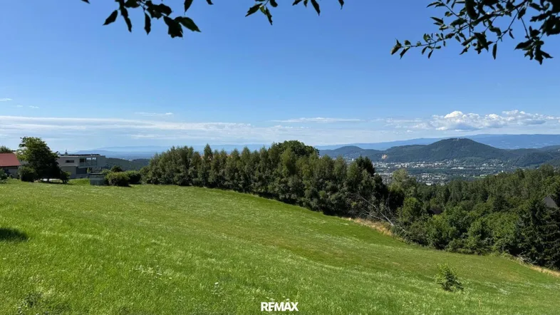 Weitläufige grüne Wiese mit Panoramablick auf die umliegende Landschaft und ferne Berge unter blauem Himmel.