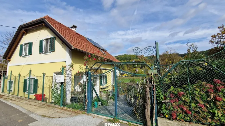 Traditionelles gelbes Haus mit roten Dachziegeln, grünen Fensterläden und einem Vorgarten.