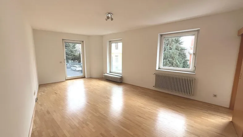 Helles, leeres Wohnzimmer mit Parkettboden, weißen Wänden und Zugang zum Außenbereich.
