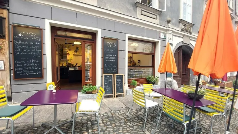 Außenbereich eines Cafés mit bunten Tischen, Stühlen und Sonnenschirmen auf Kopfsteinpflaster.