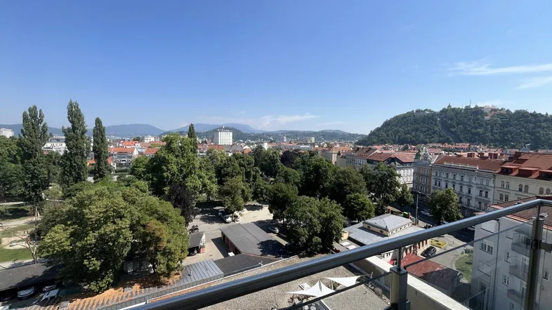 Weitläufiger Blick über die Stadt mit viel Grün und Hügeln im Hintergrund an einem sonnigen Tag.