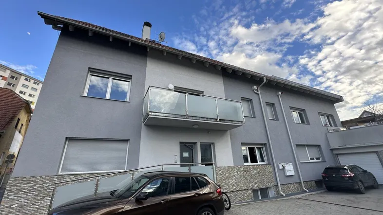 Modernes Mehrfamilienhaus mit grauer Fassade, Balkon und Parkplätzen vor dem Gebäude.