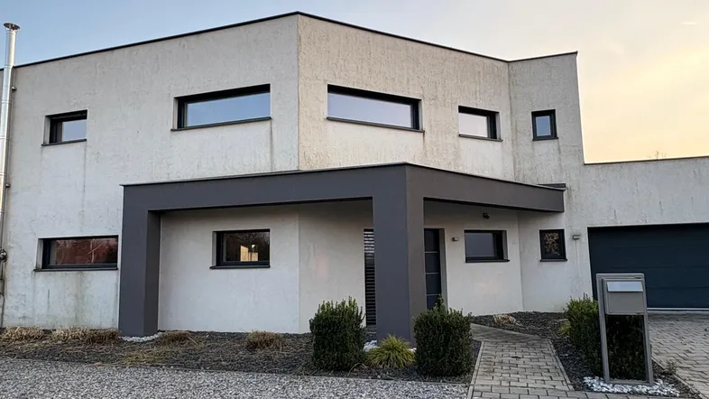 Modernes Einfamilienhaus mit heller Fassade, Flachdach und überdachtem Eingangsbereich, umgeben von gepflasterter Auffahrt und Kiesfläche.