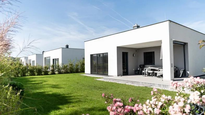 Moderne weiße Häuser mit Flachdach und gepflegtem Rasen, inklusive überdachter Terrasse mit Sitzgelegenheiten.