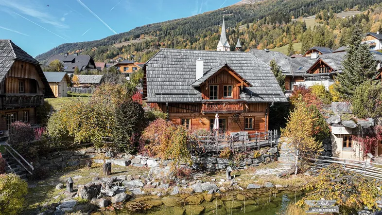 Charmantes Holzhaus in alpiner Umgebung mit Teich und Blick auf die umliegenden Berge und das Dorf.