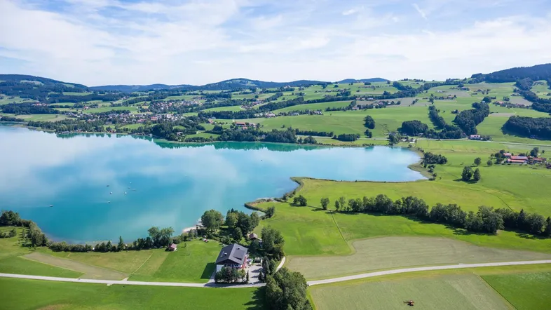 Panoramische Luftaufnahme einer weiten Landschaft mit großem See, sanften Hügeln und vereinzelten Gebäuden.